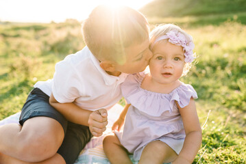 Happy children kiss and sit on blanket in green grass in field at sunset. Kids playing in mountains on summer day. Older brother plays with younger sister in spring sunlight. Spending time together.