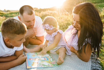 Happy young family relaxing on vacation together. Father, mother, little girl, boy reading book on blanket in green grass at sunset. Portrait of mom, dad, son, daughter. Concept of holiday on picnic.