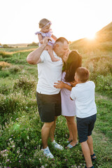 Mother kisses father, daughter girl and son boy stand in green grass in field. Children hugging parents in nature. Happy family spending time together outdoors. Family holiday at sunset on summer day