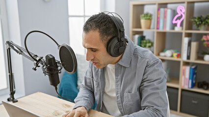 Hispanic man wearing headphones speaks into a microphone at an indoor radio studio setup,...