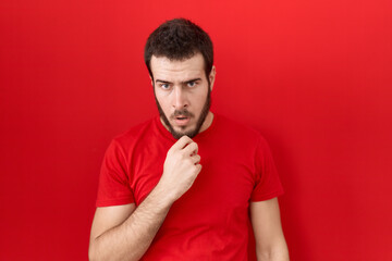 Obraz premium Young hispanic man wearing casual red t shirt looking fascinated with disbelief, surprise and amazed expression with hands on chin