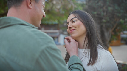 A smiling woman and a man in casual clothes share a tender moment outdoors in a lush city park.
