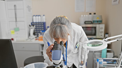 Mature woman scientist using microscope in laboratory setting, showcasing research and medical...