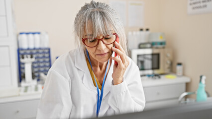 A mature woman with grey hair, wearing glasses and a lab coat, talks on the phone in a medical...