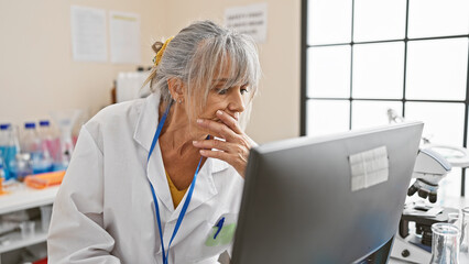 A pensive middle-aged woman in a lab coat analyzes data on a computer in a bright laboratory...