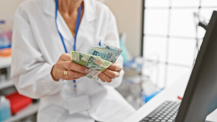 Middle-aged female doctor counts hong kong dollars indoors, highlighting healthcare costs.