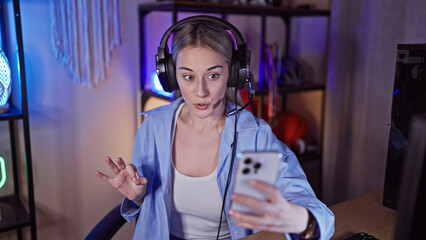 A surprised young woman with blonde hair wearing headphones looks at her smartphone in a gaming room with colorful ambient lighting.