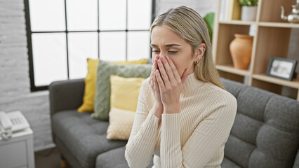 Blonde woman feels distressed sitting indoors, hand over mouth, in thoughtfully designed living...