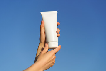 Female hands holding a white mockup tube of cream on a blue sky background. Concept of natural...