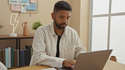 A focused young man with a beard working on a laptop in a well-organized home office setting