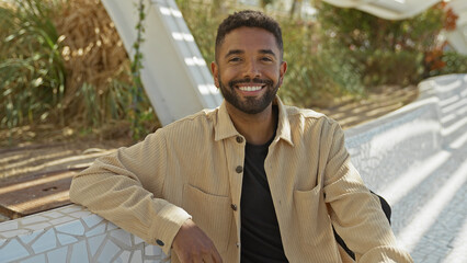 A smiling young black man with a beard wearing casual clothes sits in a city park on a sunny day.