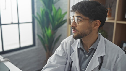 A thoughtful young man with a beard and glasses wearing medical attire in an indoor hospital setting