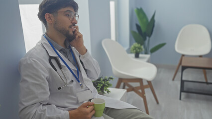 Handsome arab doctor in white coat talking on phone while holding a mug in a modern hospital...