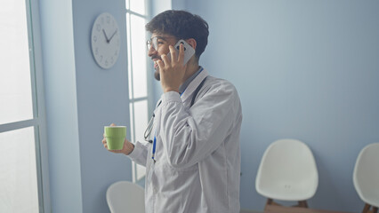 Bearded doctor in a white coat with a stethoscope talking on a smartphone indoors, holding a coffee...
