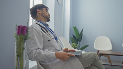 A young man with a beard, dressed as a doctor, sitting pensively in a modern hospital lobby,...