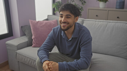 Fototapeta premium A smiling young man with a beard sitting comfortably in a modern living room apartment.