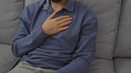 Handsome bearded man wearing blue shirt sitting on a grey couch indoors, touching chest with hand.