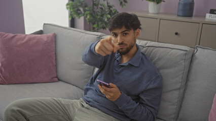 Fototapeta premium A handsome young man with a beard sitting on a couch indoors, pointing at the camera and holding a smartphone.