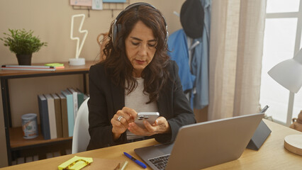 Focused middle-aged woman using smartphone and laptop in a modern home office, representing remote work.