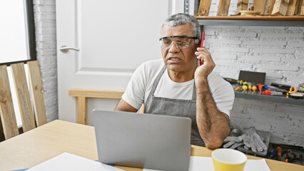 A mature man talks on the phone in his workshop with tools, glasses, and a laptop.