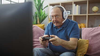 Mature man with headphones intently playing video games in a cozy living room, capturing leisure...