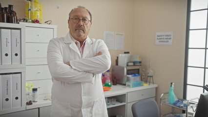 Naklejka premium Confident mature man in lab coat standing with arms crossed in a well-equipped laboratory setting