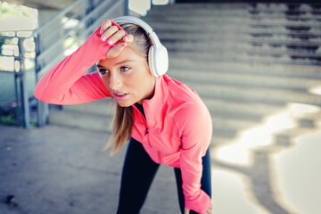 Portrait of athletic young woman resting after jogging. Beautiful blonde woman running at the city on a sunny day. Female runner listening to music while jogging.