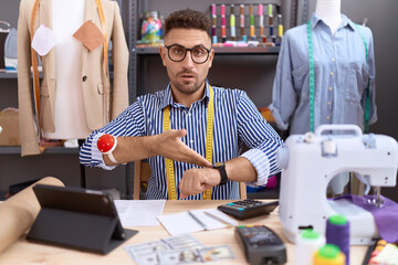 Hispanic man with beard dressmaker designer working at atelier in hurry pointing to watch time,...