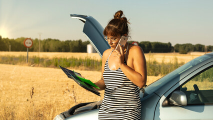 Woman looking at car insurance while talking on the phone leaning on a broken down car in the...
