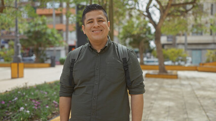 A cheerful hispanic man with a backpack standing in a city park during daytime