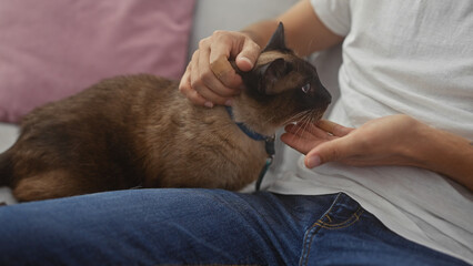 A man in a casual setting indoors lovingly pets his siamese cat sitting on his lap, exemplifying companionship and relaxation at home.