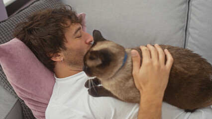 A young hispanic man affectionately pets a siamese cat while relaxing on a couch in a cozy...
