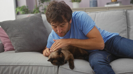 Young hispanic man affectionately pets a siamese cat while relaxing on a couch indoors.