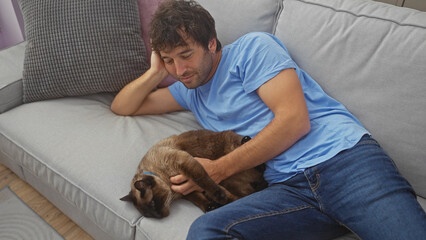 Hispanic man relaxes with pet cat on living room couch, showing bonding, comfort and leisure...