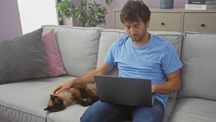 Hispanic man petting cat while working on laptop in a cozy modern living room.