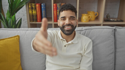 A cheerful young hispanic man with a beard extends a greeting at home, showcasing a cozy living...