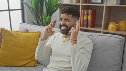 A distressed young hispanic man at home crossing his fingers for good luck while sitting on a gray...