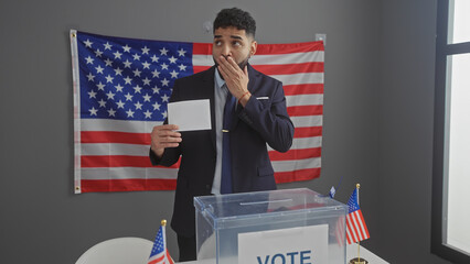 Surprised young man with a beard stands in a voting booth with an american flag in the background.