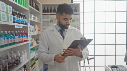 A professional hispanic man with a beard reviews a clipboard in a well-stocked pharmacy.