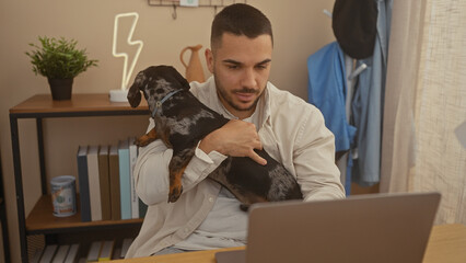 A handsome young hispanic man holding a dachshund pet dog while working on a laptop in a cozy...
