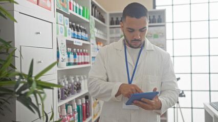 A focused hispanic man in a white lab coat uses a tablet inside a bright pharmacy full of products.