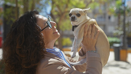 A middle-aged woman happily holding her pet chihuahua in an urban park with trees and sunlight.