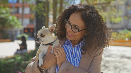 A middle-aged hispanic woman holds a chihuahua in an urban park with trees and buildings warmly...