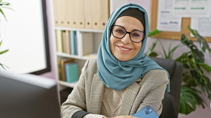 Smiling woman in hijab with glasses working in a modern office environment.