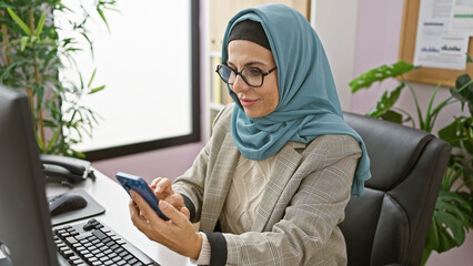 A professional woman wearing a hijab examines her smartphone in a modern office setting.