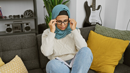 A mature woman in a hijab intently reads a book while adjusting her glasses, seated on a couch indoors.