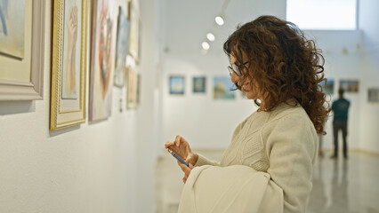 A mature woman examines art in a gallery while holding a smartphone, suggesting a blend of culture...