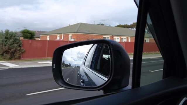 Close-up of a side-view mirror of a moving car with a blurry view of a row of suburban houses on the roadside. Concept of driving on a main road with multiple lanes in an Australian neighborhood.