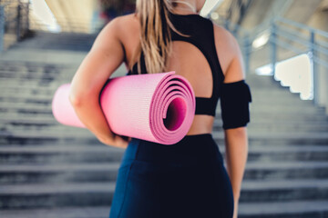 A fitness girl is standing outside on the steps, holding a yoga mat, listening to music and getting ready for her workout.