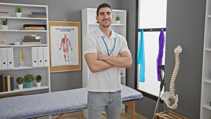 Smiling young hispanic man with crossed arms standing in a physical therapy clinic room with...